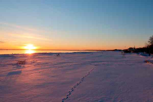 Pohled z výšky na dlouhou zasněženou pláž, se sluncem na horizontu, jeho žluto-červeným odrazem na vodě i na sněhu. Jen pár stop a pár lidí v dálce.