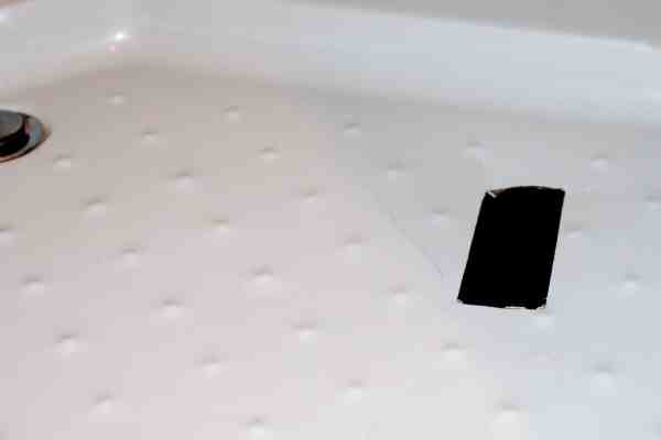 Fotodetail podlahy sprchy – vlevo je vidět odtok, v pravé polovině pak pruh černé lepící pásky. Vlevo od ní se šikmo táhne stejně dlouhá prasklina, další dvě praskliny pokračují zpod pásky.